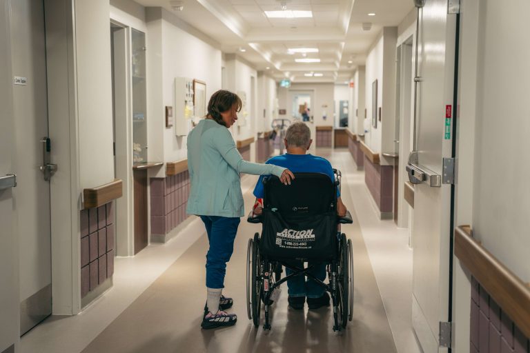 A staff member accompanying a resident in a wheelchair down a hallway