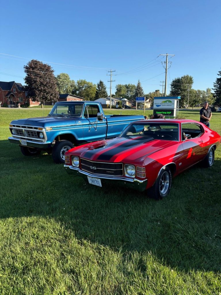 A blue Ford pickup and a red Chevelle SS parked on the lawn beside the West Perth Village sign