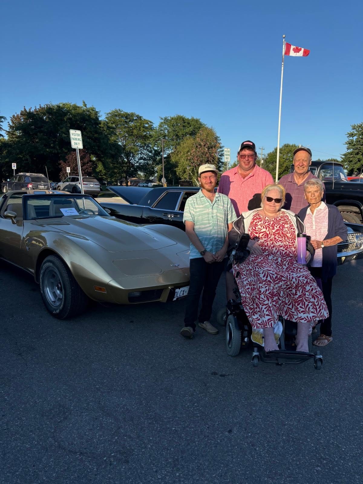 Residents and visitors beside a gold Corvette, with the Canadian flag flying behind